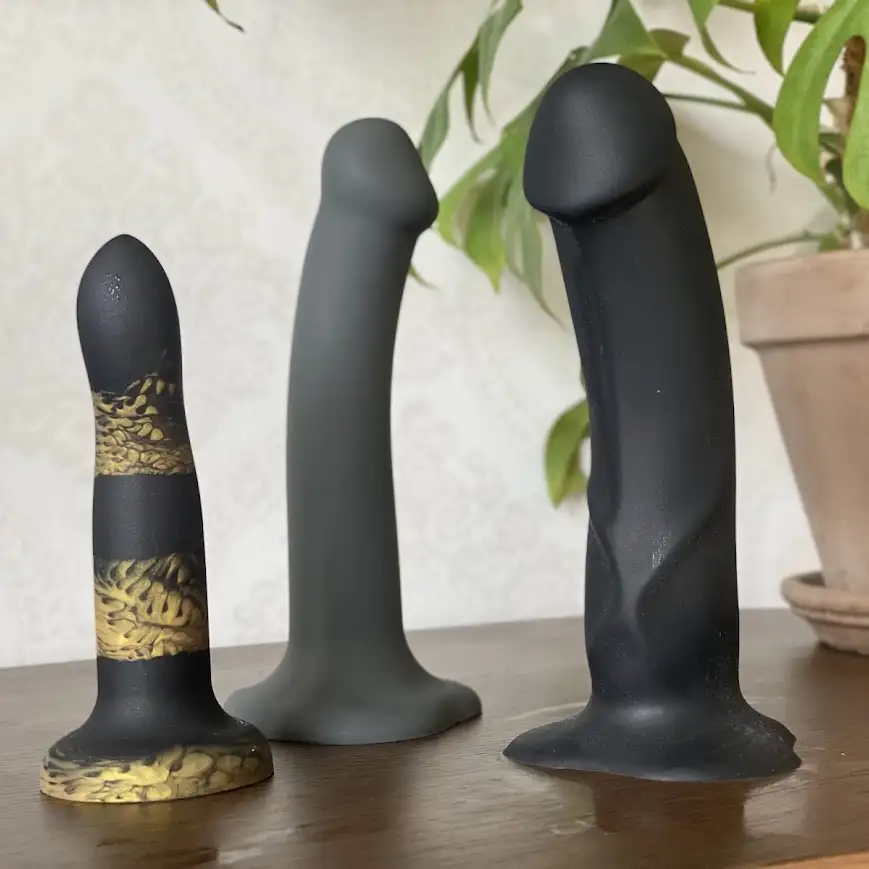 En liten tigrerad dildo för analsex, en mellanstor grå dildo och en stor dildo med ådring mot en ljus bakgrund med en växt till höger